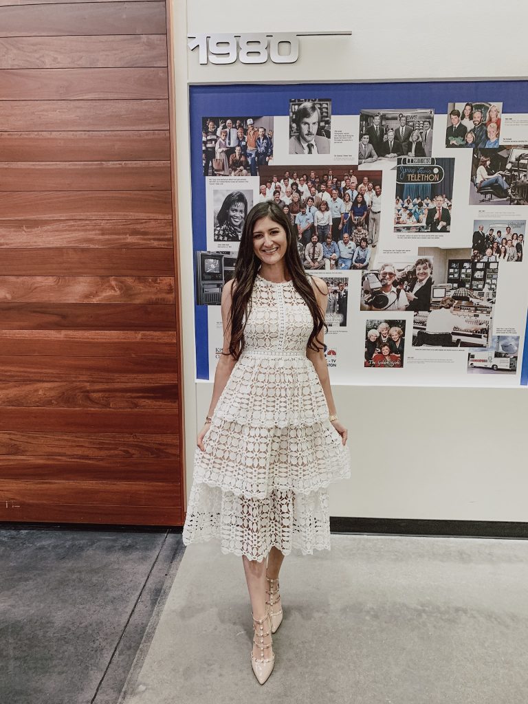 Jenni Metz standing in front of the KPRC channel 2's wall of history. She is wearing a 3 tier white crochet dress and nude studded heels. Houston life segment.