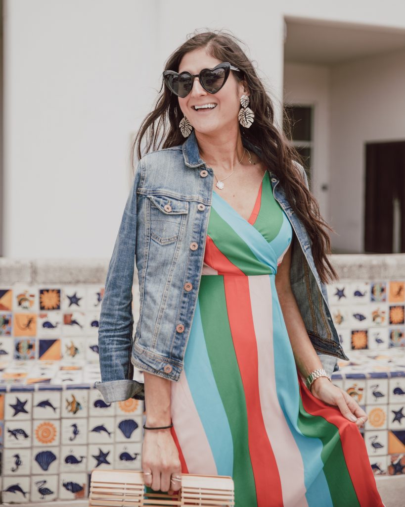 Jenni Metz- The Fashionable Maven is wearing a colorful striped dress, denim jacket, Tory Burch sandals, heart sunglasses, and bamboo bag. Click to see my round up of more striped dress outfits. #stripes