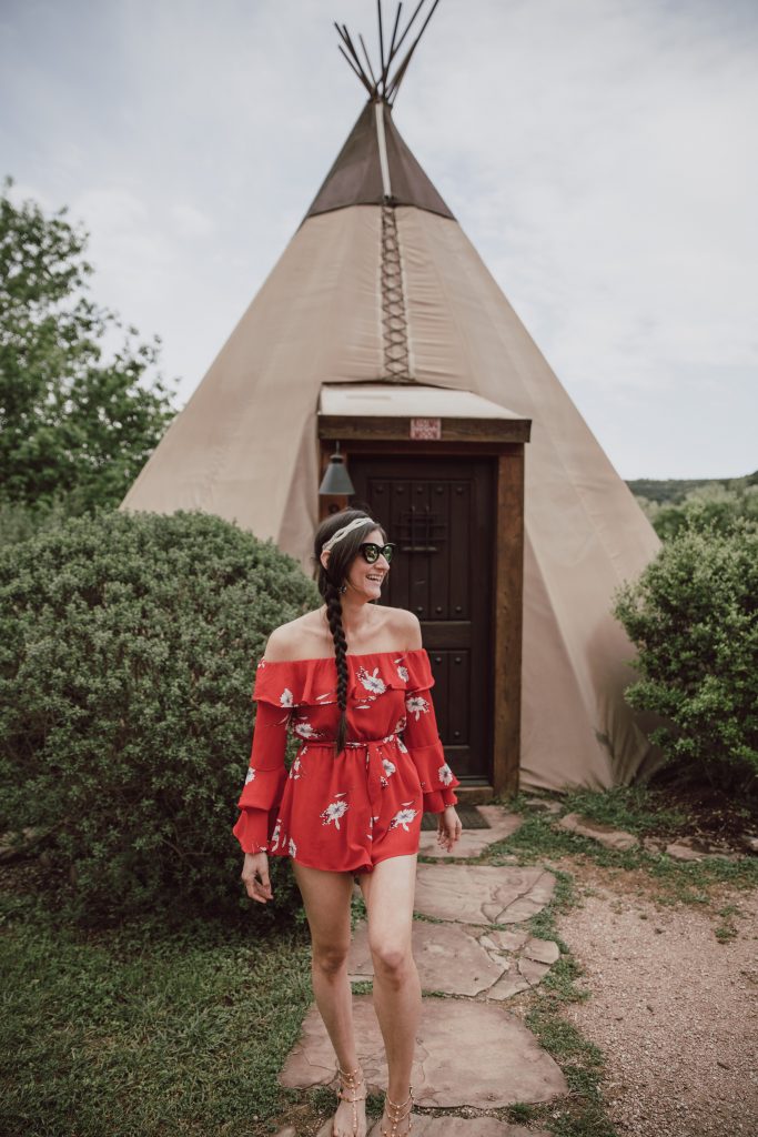 JenniMetz posing in front of the tipi camping tent. She is wearing a red off the shoulder romper.
